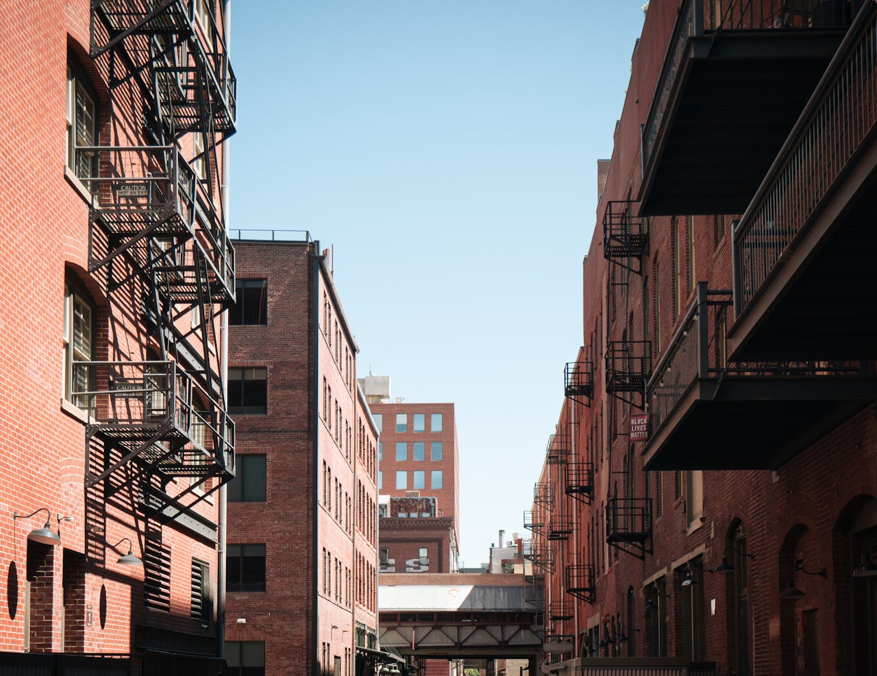 Narrow alleyway in a city with red brick buildings and fire escapes in daylight.